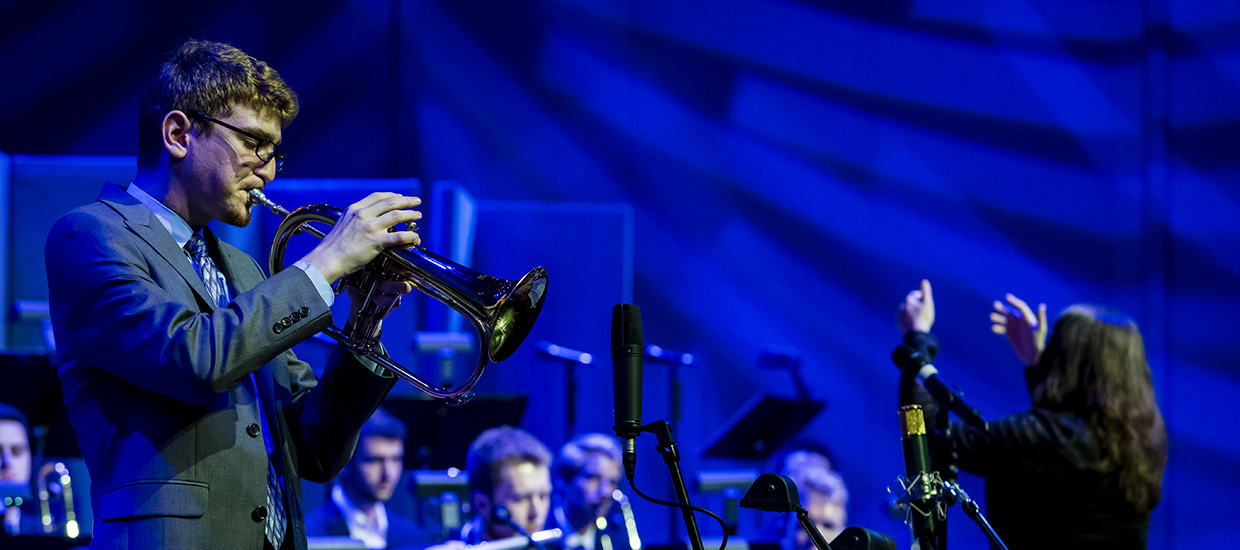 A trumpet player from the FROST Studio Jazz Band is playing while a conductor directs a band in the background