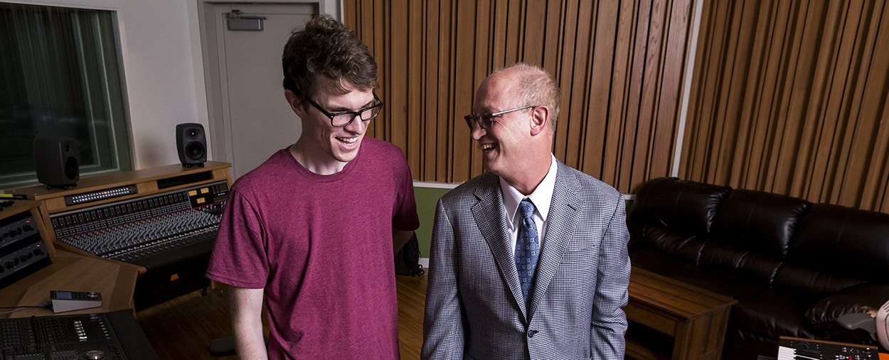 A man in a suit jacket and tie smiles at another man in a t-shirt to his right in a music studio